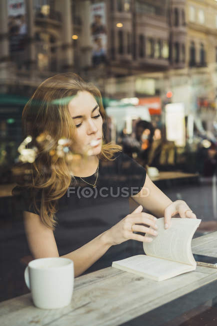 Jovem sentada com livro e café na janela do café — Fotografia de Stock