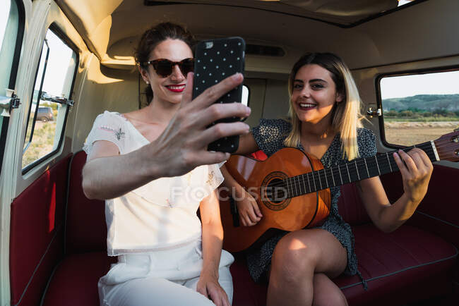 Женщины играют на акустической гитаре и смеются, сидя вместе в ретро фургоне во время поездки — стоковое фото