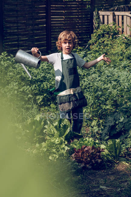 Портрет мальчика с вьющимися волосами в фартуке поливающих растений в саду — стоковое фото