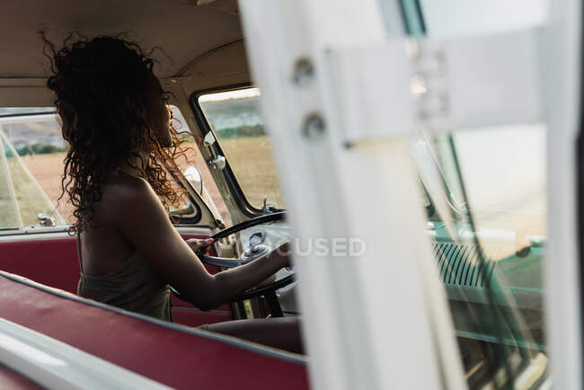Side view of charming African-American woman smiling and looking at camera while sitting on driver seat of retro car and touching curly hair during trip in countryside — Stock Photo