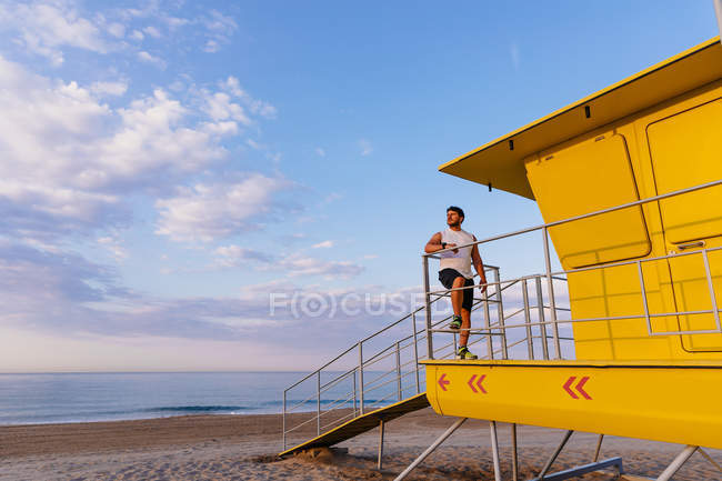 Бородатий чоловік у спортивному одязі спирається на перила рятувальної кабіни на пляжі під час тренування на свіжому повітрі на заході сонця — стокове фото