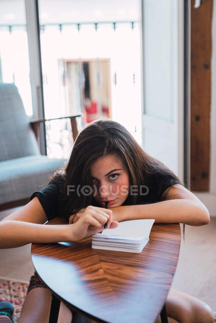 Joven mujer pensativa sentada en la pequeña mesa de madera en la habitación y la escritura en el diario - foto de stock