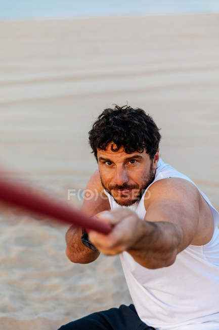Hombre barbudo seguro en ropa deportiva tirando de la cuerda mientras hace ejercicio en la playa de arena - foto de stock