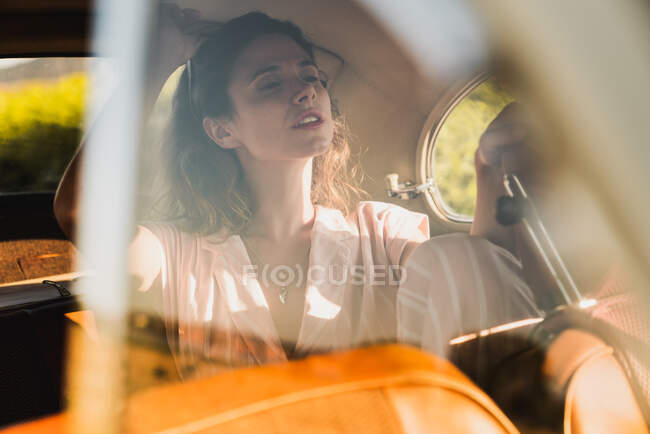 Уверенная женщина в розовых очках и солнцезащитных очках, сидящая на заднем сиденье в ретро-машине и отводящая взгляд от солнца — стоковое фото