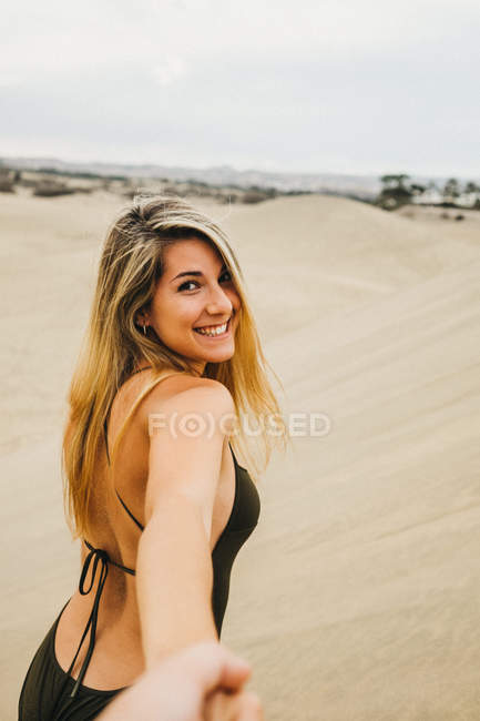 Fröhliche junge Frau im Badeanzug lächelt und blickt in die Kamera, während sie am Sandstrand den Weg weist — Stockfoto