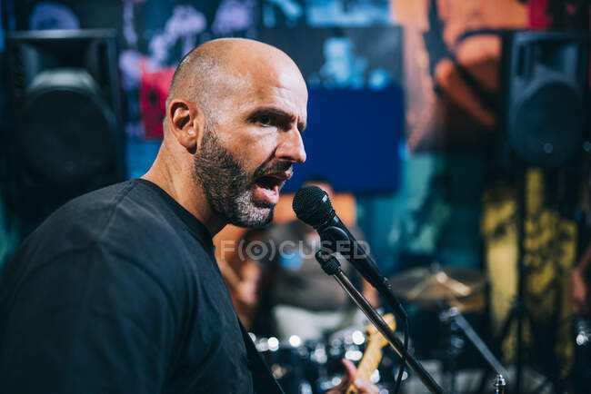 Vista lateral del hombre calvo en camiseta negra de pie cerca del micrófono en el escenario y cantando sobre fondo borroso - foto de stock