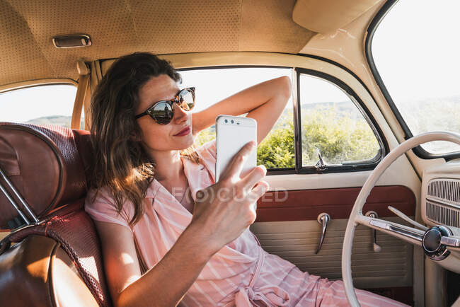 Hermosa morena en vestido rosa y gafas de sol de moda usando el teléfono y tomando selfie dentro de coche retro - foto de stock