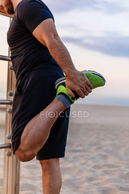 Unrecognizable muscular guy in sportswear doing warming up exercise for legs while standing near ladder during sunset on beach — Stock Photo