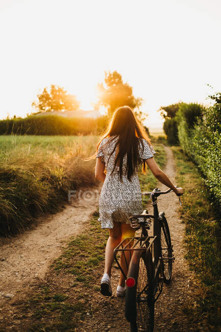 Задний вид на красивую женщину с велосипедом и прогулки по сельской дороге в подсветке — стоковое фото