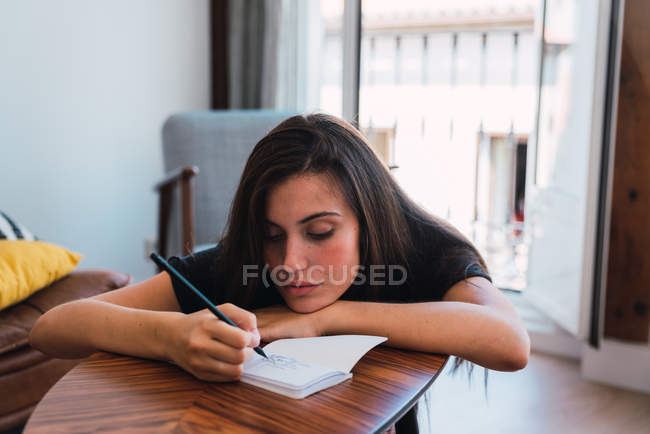 Young thoughtful woman sitting at wooden small table in room and writing in diary — Stock Photo