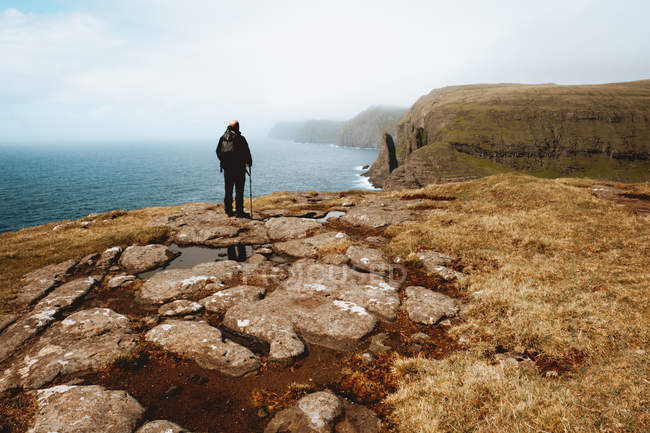 Uomo escursionista irriconoscibile in piedi sulla costa con bastone e guardando l'oceano sulle isole Feroe — Foto stock
