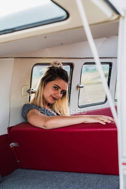 Belle femme assise sur le siège arrière confortable du van vintage et regardant loin tout en voyageant dans la campagne — Photo de stock