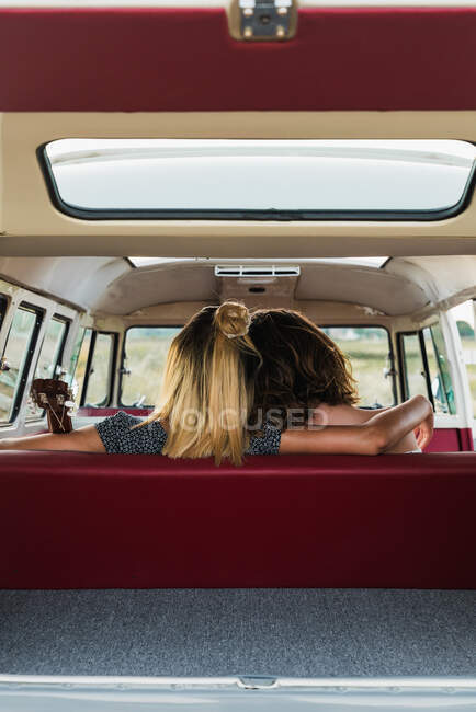 Vista trasera de dos mujeres abrazándose mientras están sentadas en el asiento trasero de la furgoneta retro en la naturaleza - foto de stock