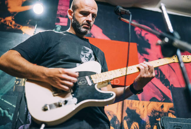 De dessous vue de l'homme chauve en T-shirt noir jouant de la guitare électrique sur scène sur fond de photos — Photo de stock