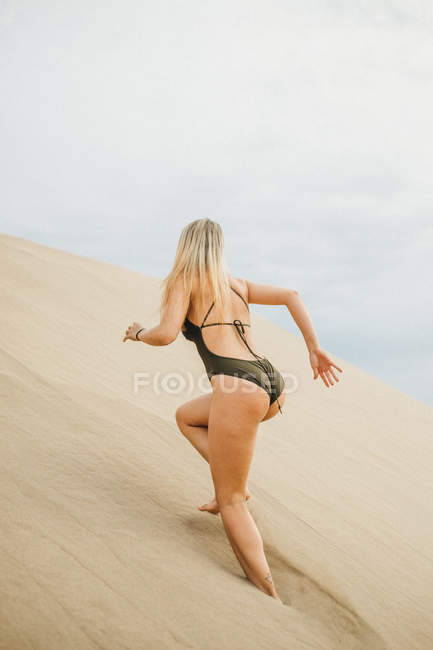 Mujer rubia joven en traje de baño negro corriendo hasta la duna de arena mientras descansa en la playa - foto de stock
