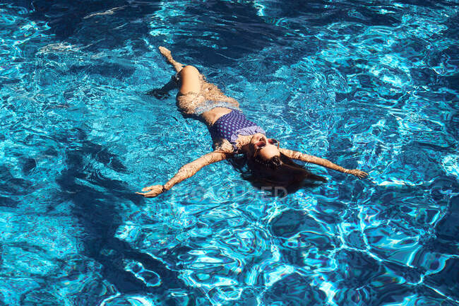 Зверху молода брюнетка в купальнику і сонцезахисних окулярах відпочиває на блакитній чистій воді басейну — стокове фото