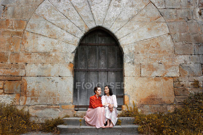 Senhoras multiétnicas atraentes sentadas em degraus perto da porta da torre e olhando para longe — Fotografia de Stock