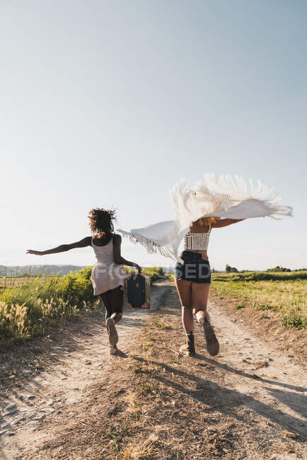 Mulheres alegres e elegantes multiétnicas com mala e cachecol correndo excitadamente na estrada no campo verde de verão — Fotografia de Stock