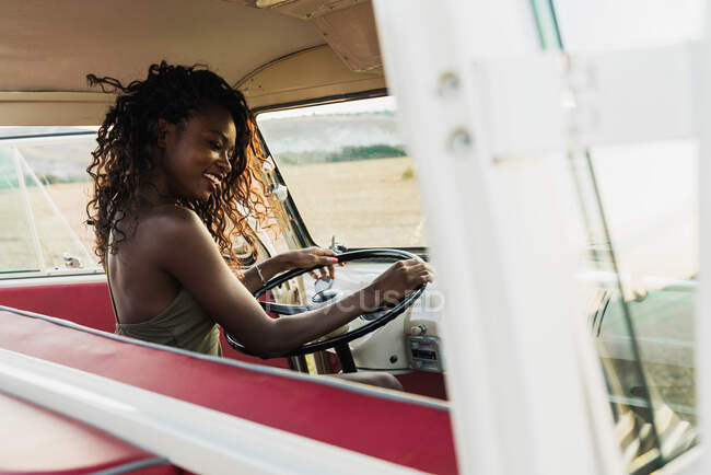 Side view of charming African-American woman smiling and looking at camera while sitting on driver seat of retro car and touching curly hair during trip in countryside — Stock Photo