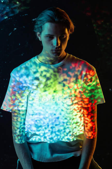 Androgynous male model looking on bright specks of red and blue light on T-shirt while standing on black background — Stock Photo