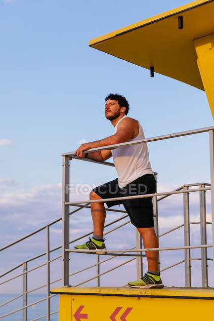 Hombre barbudo en ropa deportiva apoyado en la barandilla de la cabina de salvavidas en la playa durante el entrenamiento al aire libre al atardecer - foto de stock