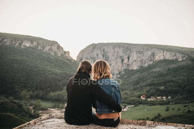 Милая пара обнимается, сидя на скалистом склоне на фоне красивой долины и гор — стоковое фото