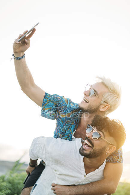Handsome bearded guy giving piggyback ride to cheerful boyfriend and posing for selfie while spending time in nature together — Stock Photo