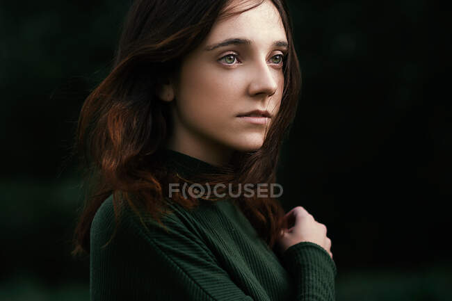 Pretty young lady in green turtleneck shirt touching shoulder and looking away while standing on dark background of nature — Stock Photo
