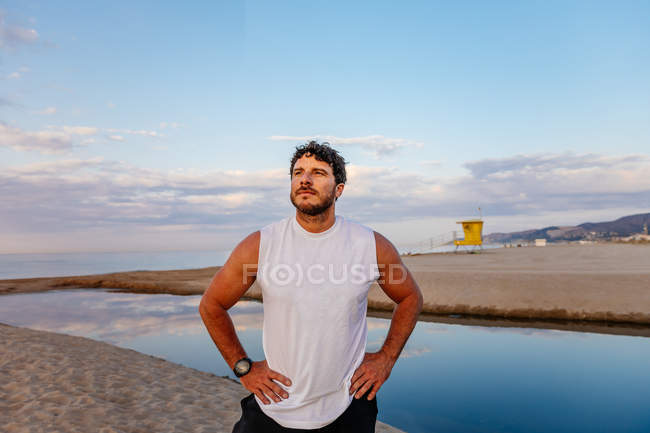 Позитивный мужчина в спортивной одежде с руками на талии, стоя на песчаном пляже во время заката — стоковое фото