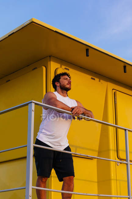 Bearded man in sportswear leaning on railing of lifeguard cabin on beach at sunset — Stock Photo