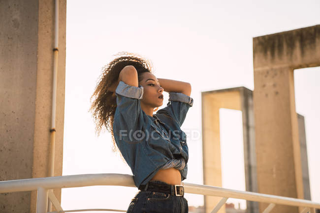 Young woman in denim clothes standing at balcony railing and touching hair — Stock Photo