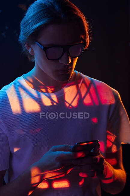 Schöner androgyner Mann mit stylischer Sonnenbrille, der im dunklen Raum unter knallroter Beleuchtung im modernen Smartphone surft — Stockfoto