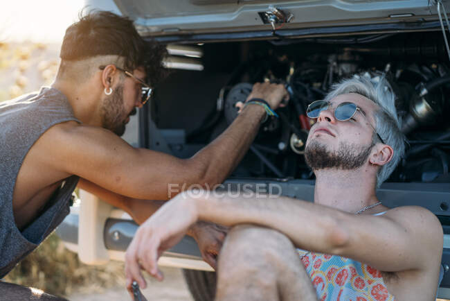 Young male leaning back and relaxing while sitting near friend trying to repair broken motor of car while during travel in nature — Stock Photo