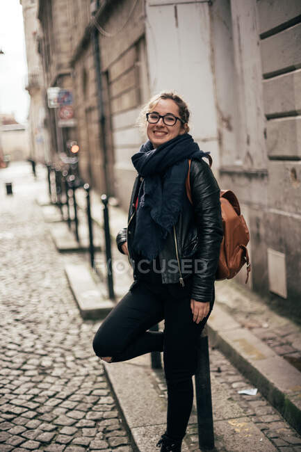 Vista lateral da linda jovem fêmea em roupa casual sorrindo e olhando para a câmera enquanto se inclina no pólo na rua da incrível cidade velha — Fotografia de Stock