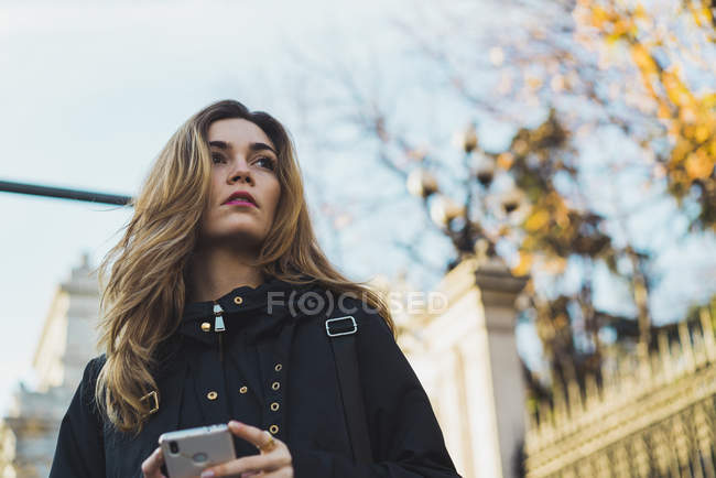 Молодая женщина держит смартфон в городе и смотрит в сторону — стоковое фото