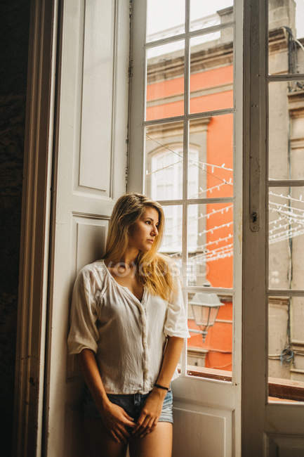 Портрет молодой женщины в повседневной одежде, смотрящей в окно в уютной комнате — стоковое фото