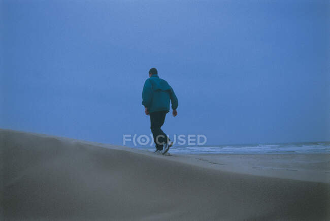 Задний вид человека в теплой куртке и брюках прогуливаясь по песчаному пляжу у моря на фоне голубого безоблачного неба — стоковое фото