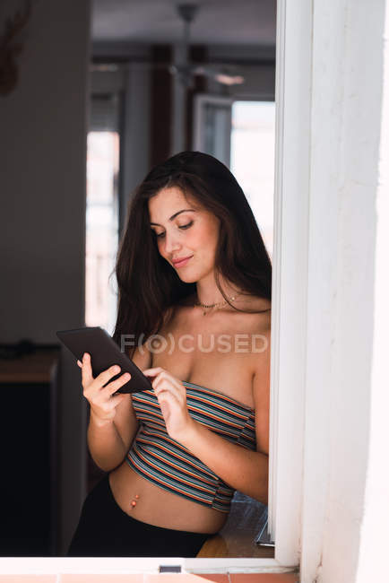 Jovem mulher pensativa de pé perto da janela e usando telefone celular — Fotografia de Stock