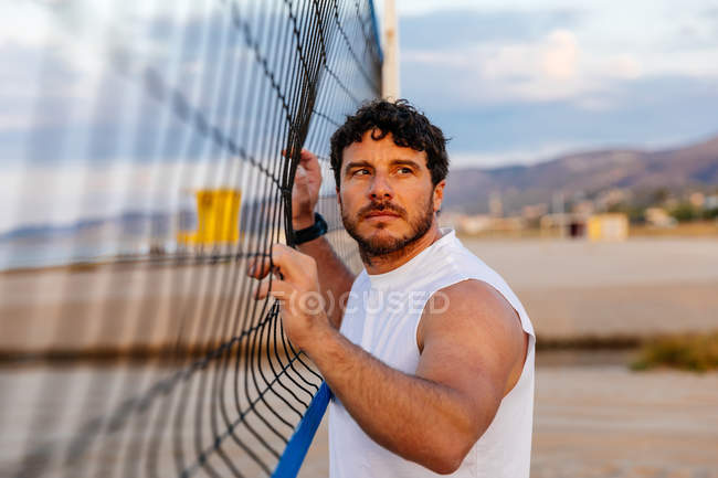 Bearded man in sportswear touching volleyball net and looking away during training on beach at sunset — Stock Photo