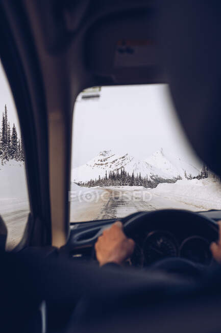 Male holding helm in dark interior and driving vehicle on road passing through forest on white snowy Canadian mountains background in soft focus — Stock Photo