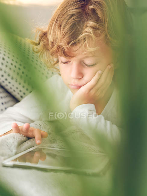 Набридлий хлопчик натискає екран цифрових планшетів, лежачи на зручному дивані — стокове фото