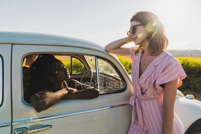 Stylish brunette in sunglasses leaning on car outside and talking to pretty black woman inside in summer bright sunlight — Stock Photo