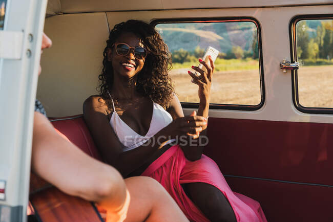 Разнообразные женщины, играющие на акустической гитаре и смеющиеся, сидя вместе в ретро фургоне во время поездки — стоковое фото