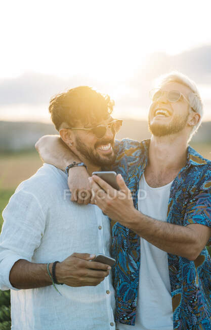 Dos hombres sonrientes abrazando y navegando teléfonos inteligentes mientras están de pie en un hermoso campo juntos - foto de stock