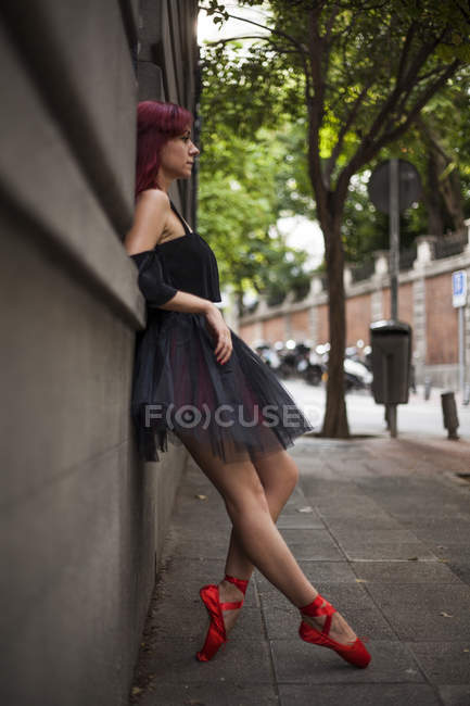 Bailarina pelirroja con tutú negro y puntas de ballet rojo calentándose para bailar en la calle, realizando poses, estampadas en la pared - foto de stock