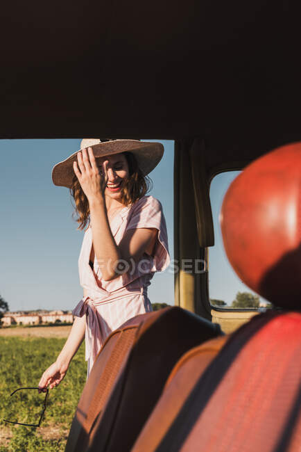 View from inside of retro car of happy trendy woman in hat and sunglasses standing excitedly in nature and laughing — Stock Photo