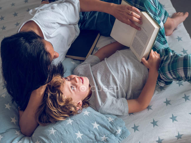 З верхнього кадру милого хлопчика лежить на гарному ліжку і читає цікаву книжку з допомогою матері. — стокове фото