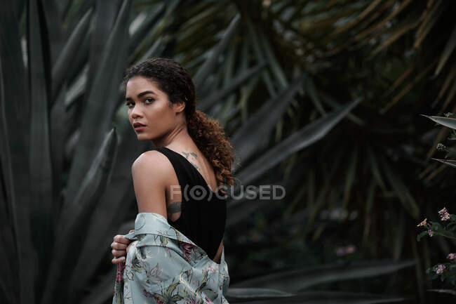 Visão traseira da bela jovem afro-americana encantadora com lenço olhando para a câmera e de pé perto de plantas — Fotografia de Stock