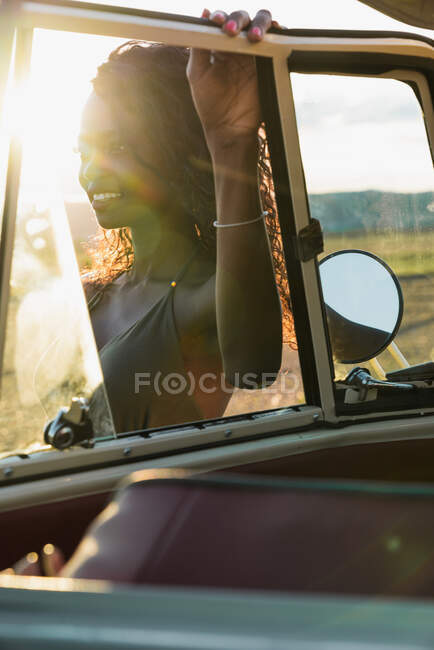 Preciosa mujer afroamericana sonriendo y mirando a través de la ventana del coche vintage mientras pasa tiempo en la naturaleza en el día soleado - foto de stock