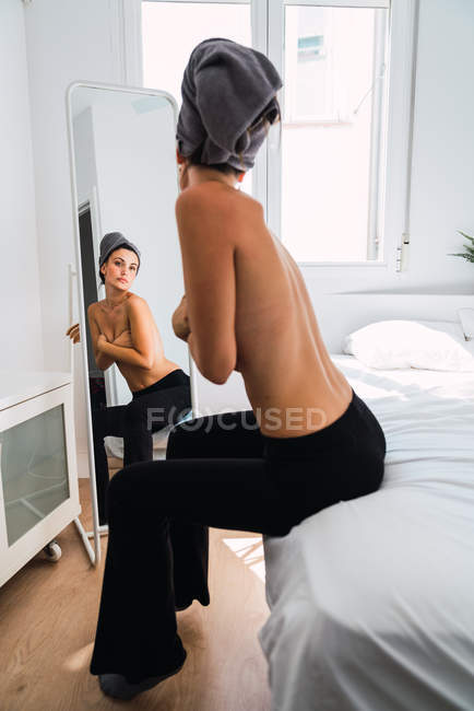 Молода топлес жінка в чорних штанях стоїть перед дзеркалом з рушником на голові — стокове фото